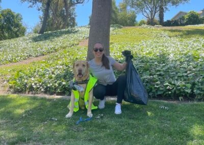 A woman and a golden retriever dog in a neon vest sit on the grass in a park, with the woman holding a trash bag