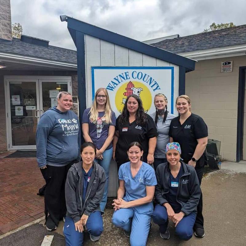 group of veterinary team members posing in front of a sign that reads Wayne County which is the part of an animal clinic logo that is visible in the photo